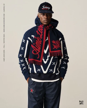 Man in a blue and white patterned sweater, blue pants, scarf, and hat stands against a neutral backdrop in a studio shot.