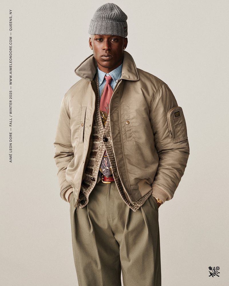 Man in tan bomber jacket, cardigan, blue shirt, tie, pleated pants, and grey beanie, posing in a studio environment.