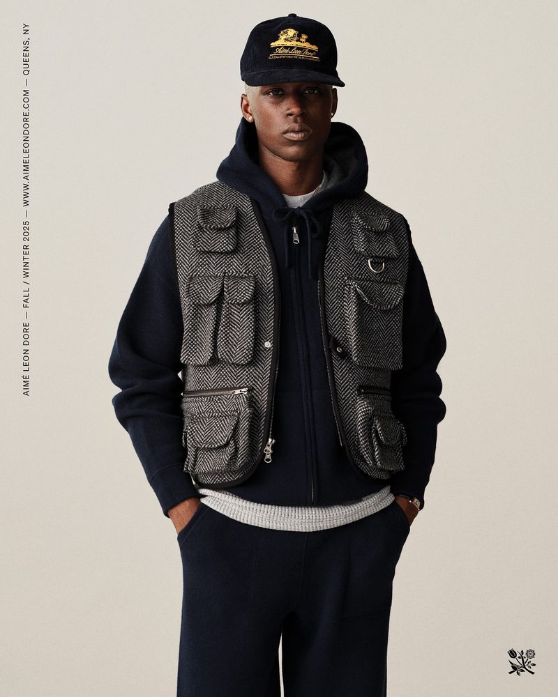 Fashion shot of a man wearing a multi-pocket vest, hooded sweater, and baseball cap, posed in a studio setting, with text on the left side.
