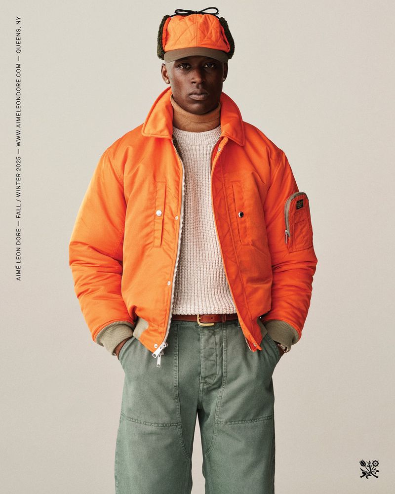 Male model poses in an orange jacket, knit sweater, and green pants, accessorized with a trapper hat against a neutral backdrop.