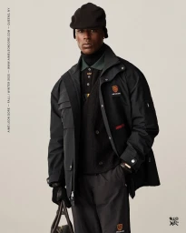Model wearing a black jacket, dark cap, and holding a bag against a solid light grey studio backdrop.