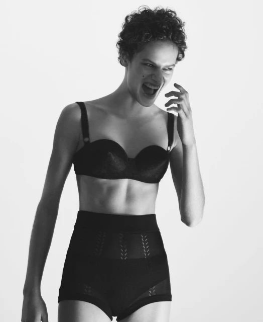 Monochromatic studio portrait of a young adult woman with curly hair, expressive face, and wearing black lingerie set against a white background.