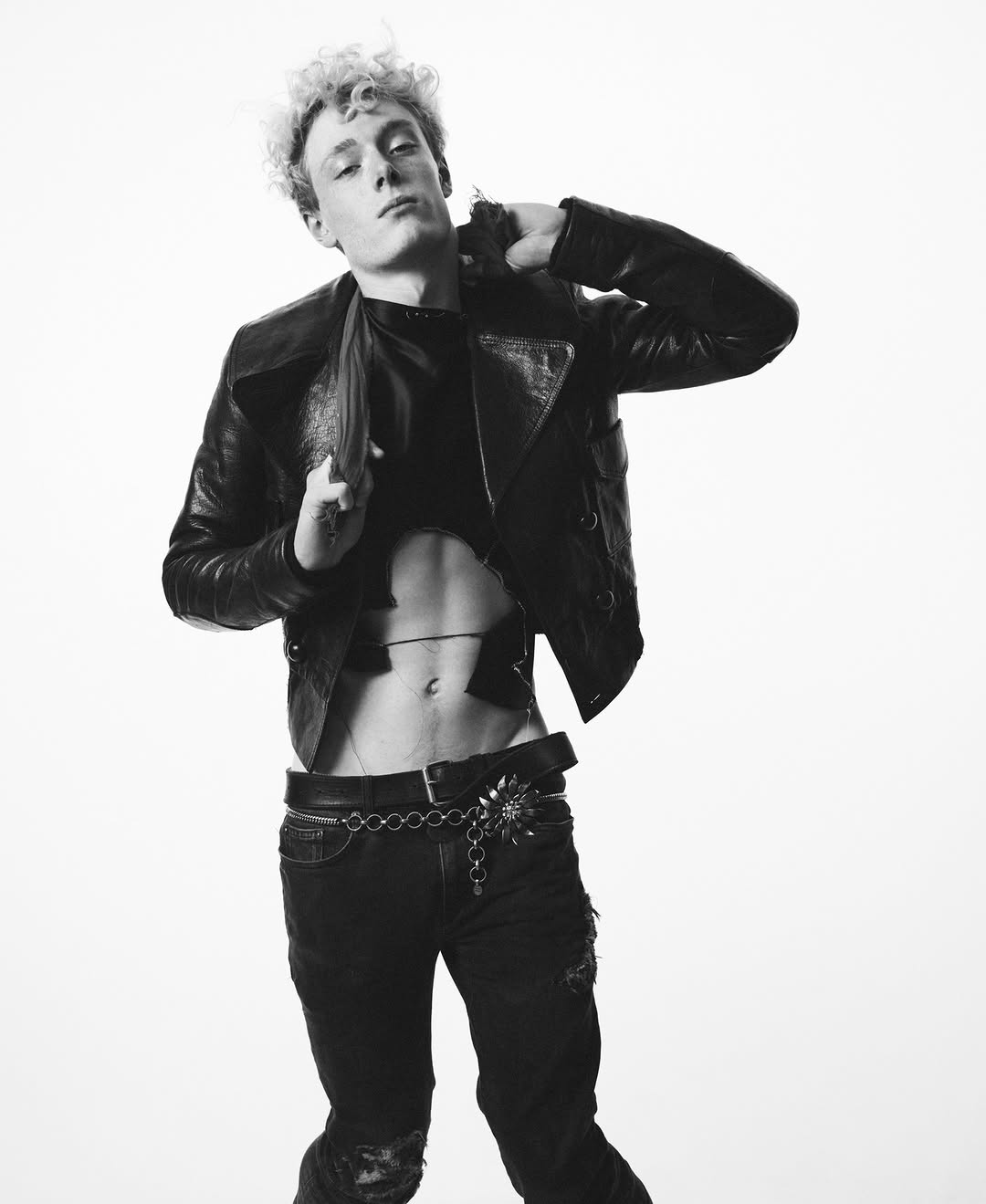 Black and white portrait of a young adult male with curly hair, wearing a leather jacket and jeans against a stark white backdrop.