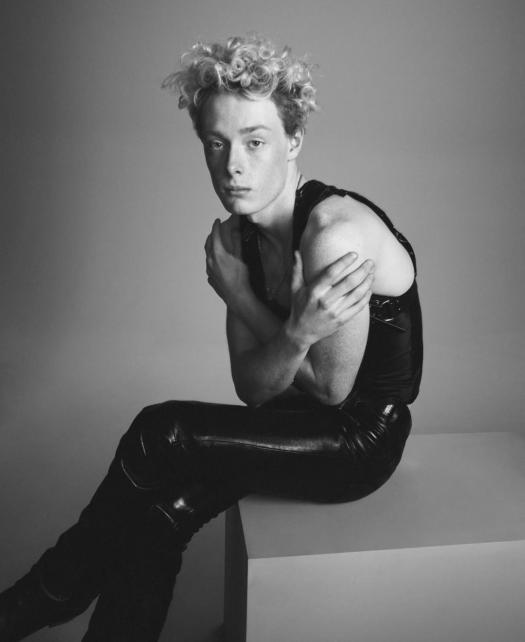 Monochrome portrait of a young adult male seated on a cube, arms crossed, wearing a sleeveless shirt and shiny pants in a photo studio.