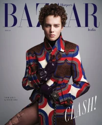Model with curly hair wearing a patterned geometric coat and dark gloves against a grey backdrop