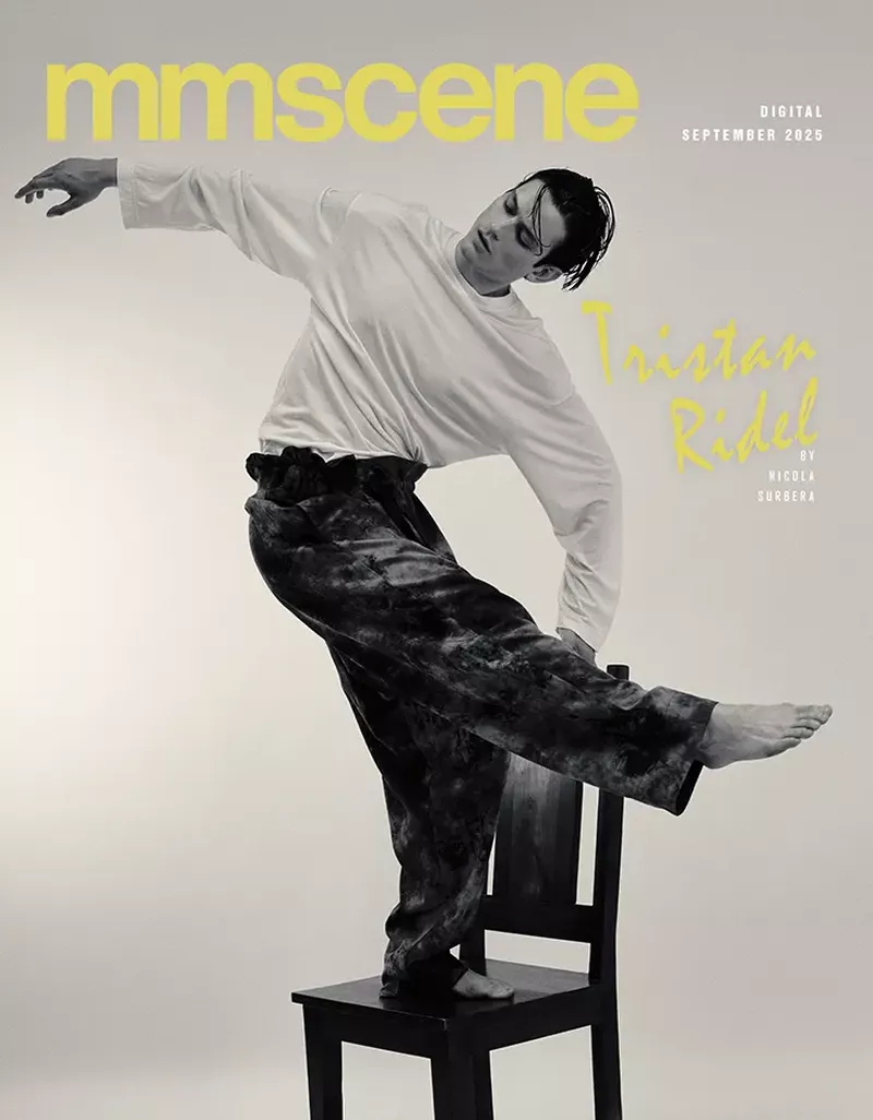 A male model in a white long-sleeve shirt and dark pants poses dynamically on a dark wooden chair in a studio setting.