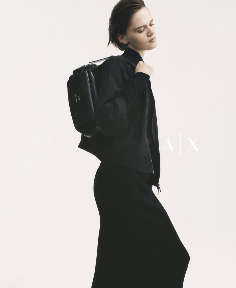A woman poses in black jacket and long skirt with a matching backpack against a white background in a fashion studio shot.