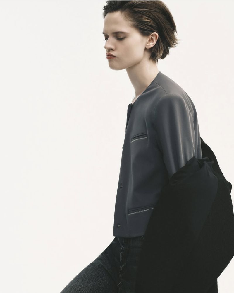 Androgynous person in a studio wearing gray blazer and dark denim jeans looking away in a neutral pose with a white backdrop.