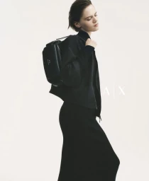 Model wearing a black jacket and carrying a black duffel bag against a white seamless background for Armani Exchange.