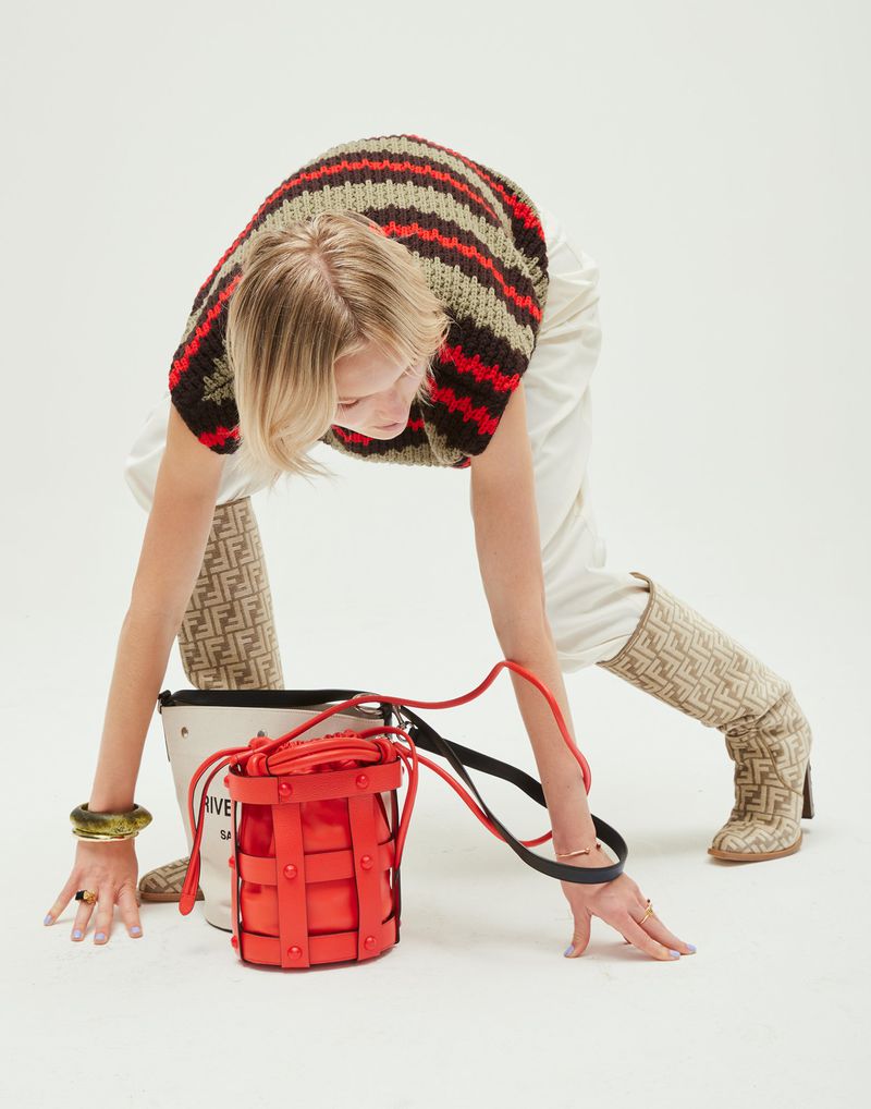 Fashion shoot featuring a young woman in a striped knit top and monogrammed boots, leaning over a red bucket bag on a white background.
