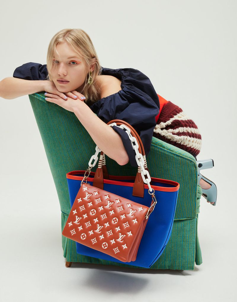 Woman poses with a stylish brown and blue tote bag with monogram design in a studio setting, paired with a blue top and green chair.