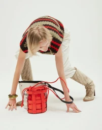 Model leaning over a red bucket bag wearing a striped knit top, white trousers, and monogram knee-high boots