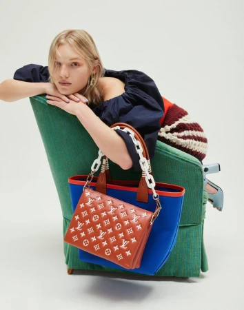 Model leaning over a green armchair holding a blue and brown designer tote bag against a seamless white background
