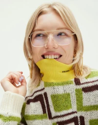 Close-up portrait of a blonde woman wearing gold-framed glasses and a green patterned sweater against a white background