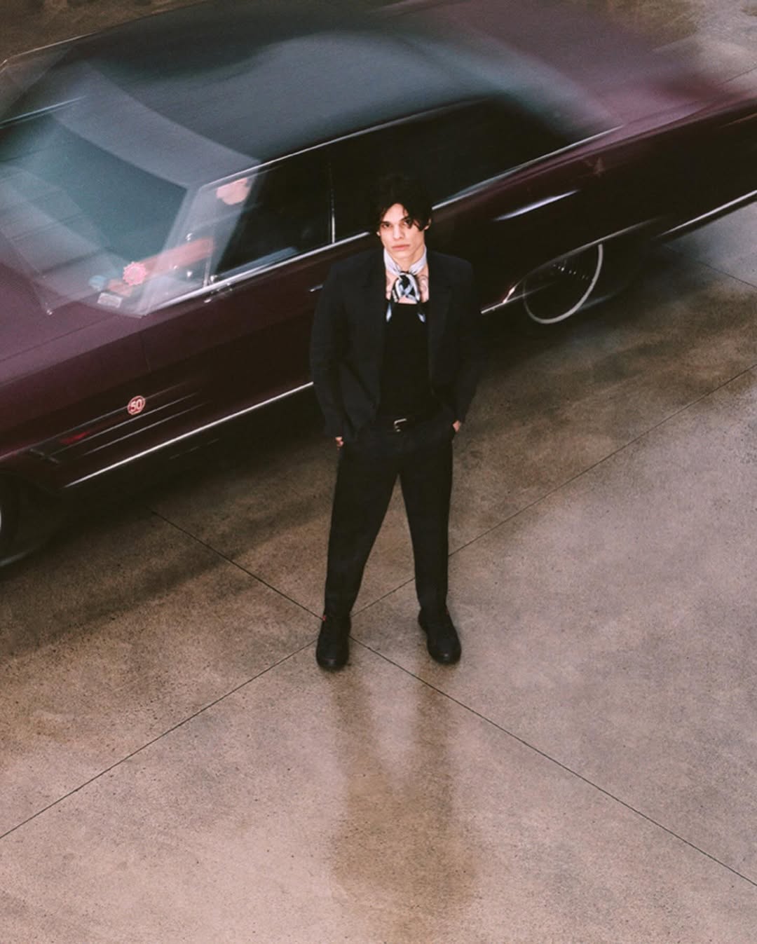 Adult male in a black suit with a cravat standing next to a dark colored vintage car on a concrete surface, medium shot.