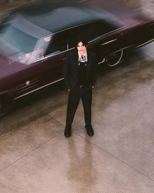 Adult male in a black suit with a cravat standing next to a dark colored vintage car on a concrete surface, medium shot.