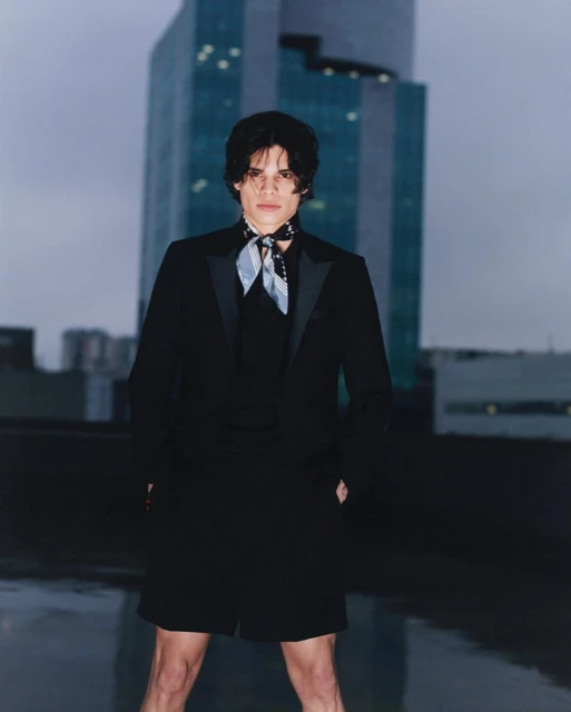 Young man in a black suit with a neck scarf stands on a rooftop in a city setting. A tall building rises in the background.