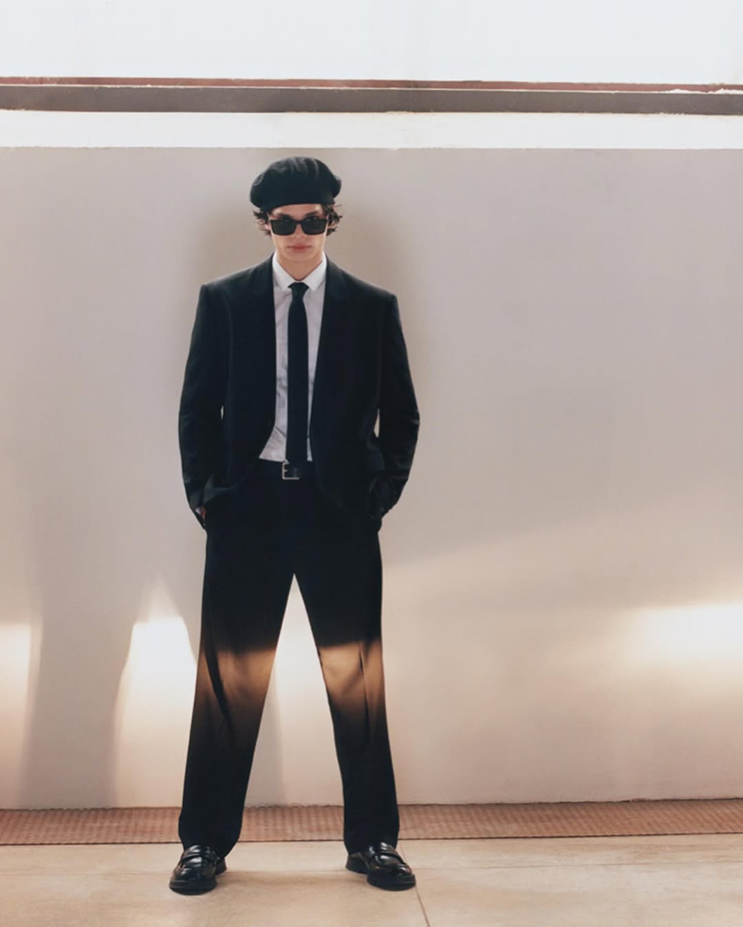 A man wearing a black suit, dark sunglasses, and a beret stands against a plain neutral background.