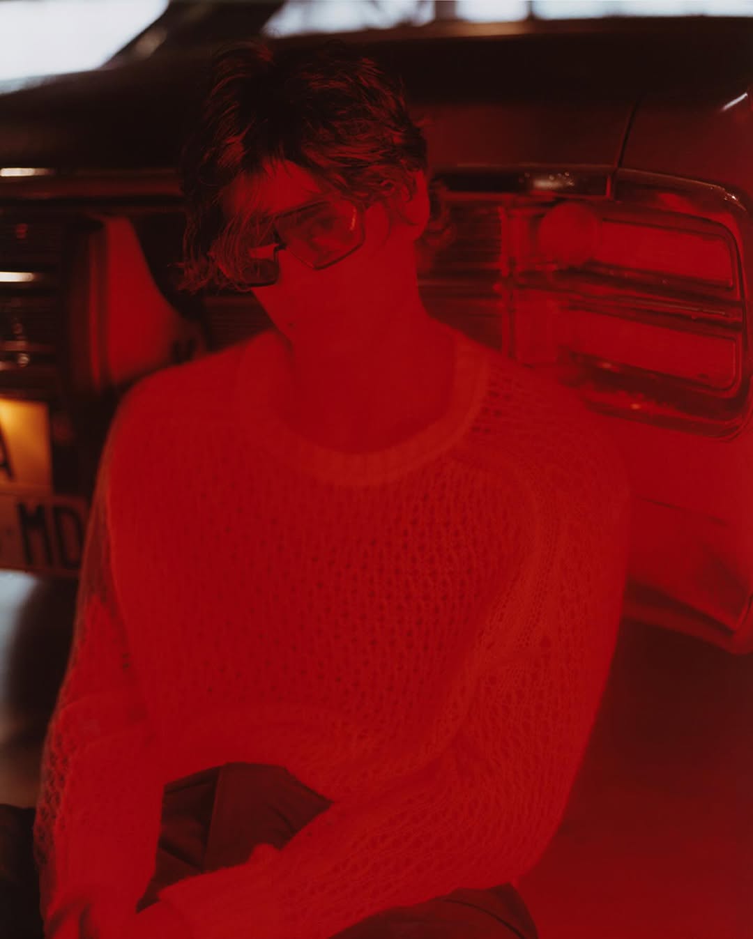 Adult in white sweater stands near a red car with a license plate in an indoor setting under red lighting. The scene is intimate and focused.
