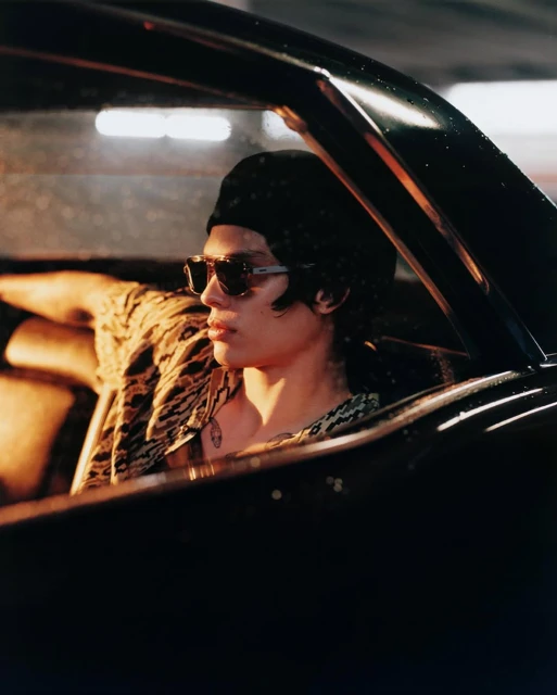 Young adult man wearing sunglasses and head covering in a black car. He is wearing an open animal print shirt and a pendant necklace.