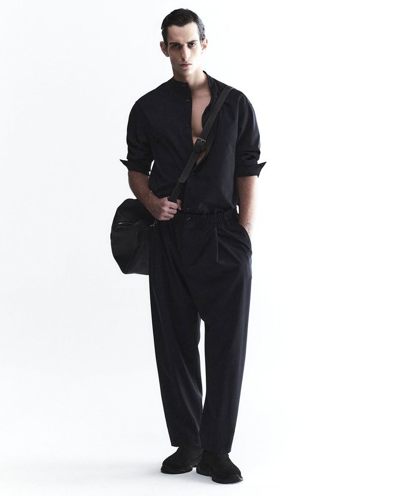 Full-length studio shot of a male fashion model in a black button-down shirt, baggy pants, and carrying a black messenger bag on a white backdrop.