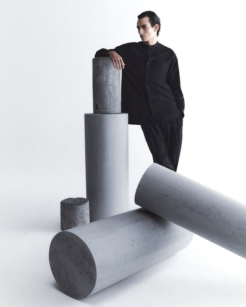 Male fashion model in black shirt poses near concrete cylinders in a studio setting, showcasing modern menswear and minimalist design.
