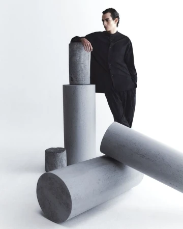 Male fashion model in black shirt poses near concrete cylinders in a studio setting, showcasing modern menswear and minimalist design.