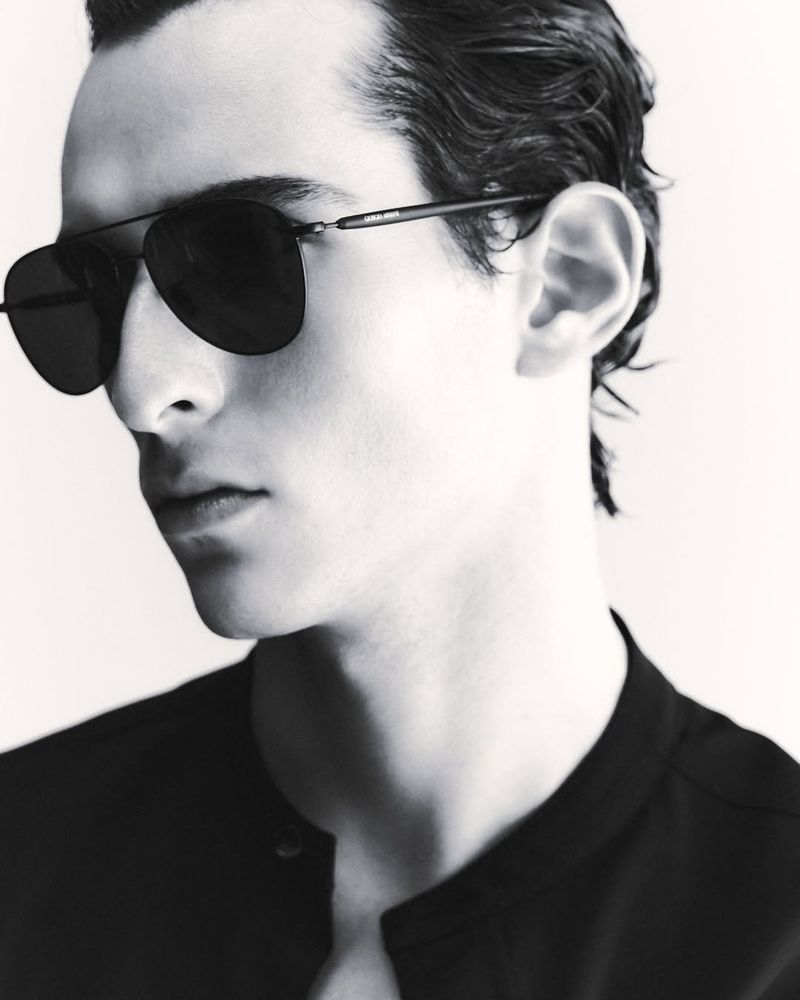 Black-and-white portrait of a young adult man with sunglasses, wearing a buttoned shirt in a bright studio setting, captured in a close-up.