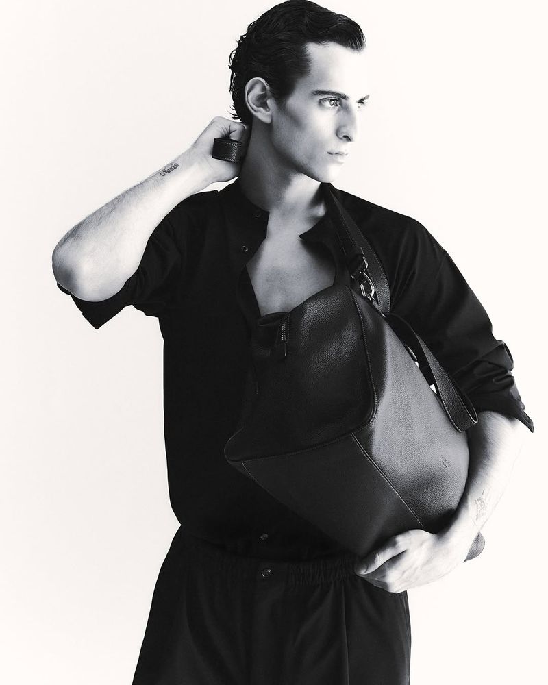Monochromatic portrait of a slender male model in a button-down shirt with a large leather bag slung over his shoulder, looking off-camera.