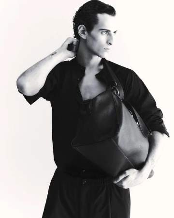 Monochromatic portrait of a slender male model in a button-down shirt with a large leather bag slung over his shoulder, looking off-camera.