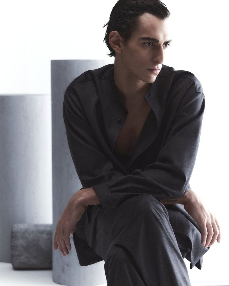 Portrait of a slender adult male in a dark button-down shirt and pants sitting in a photo studio, looking off to the side.