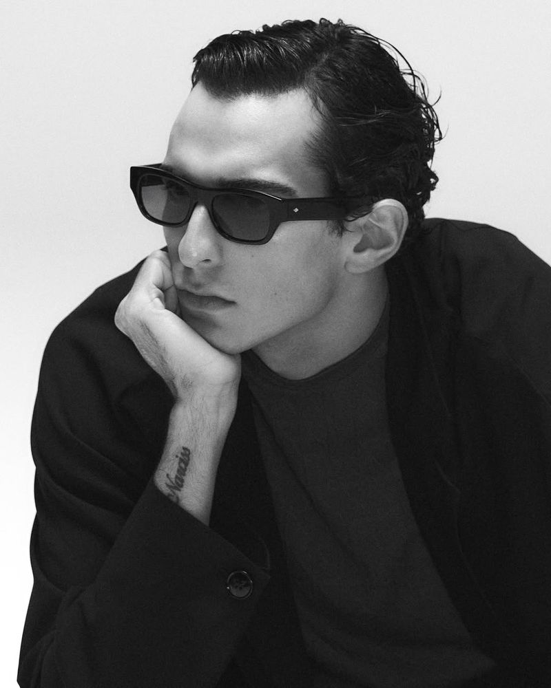 Monochrome portrait of a young adult man with slicked-back hair wearing sunglasses and a dark coat, posing against a light background.