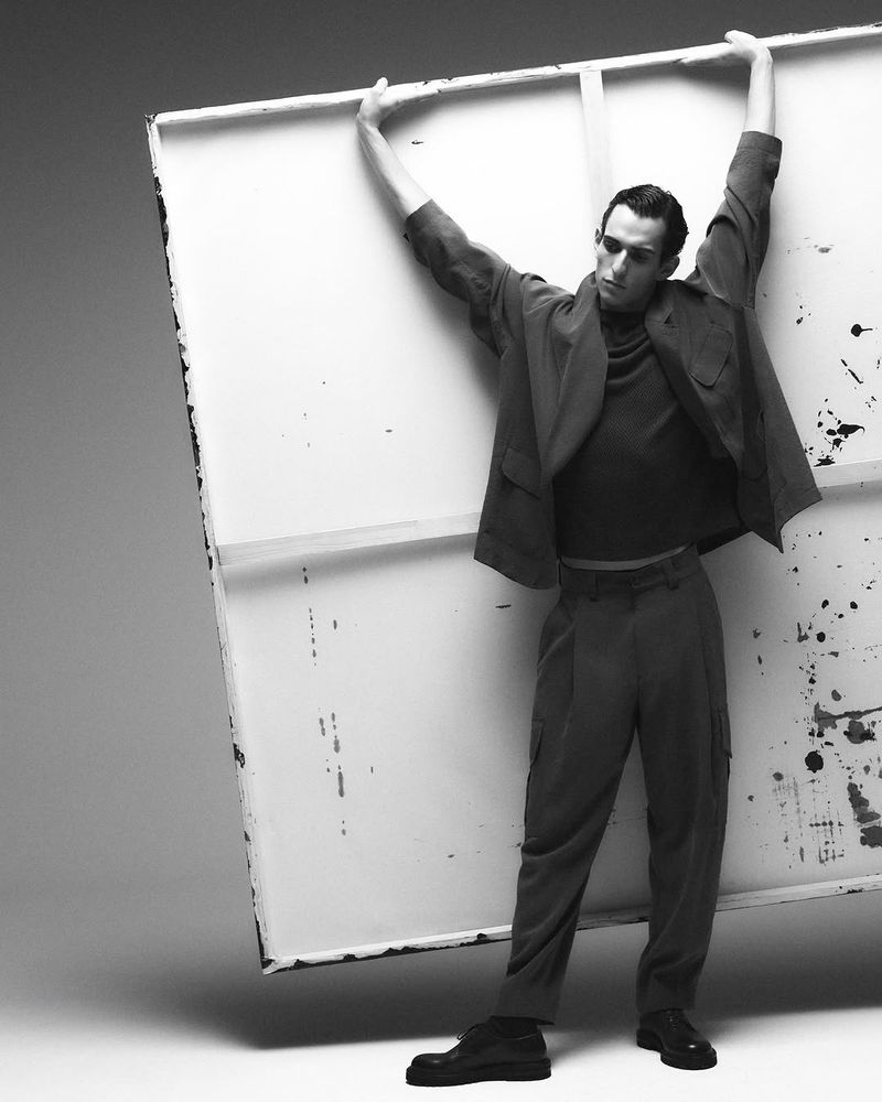 A fashionable adult male in a suit holds a white painting canvas against a gray backdrop in a studio. He is wearing dark shoes.