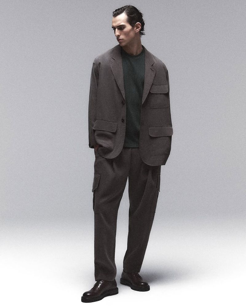 A full-length studio shot captures a slender adult male in a relaxed pose, wearing a brown blazer suit with an olive t-shirt and dark brown leather shoes.