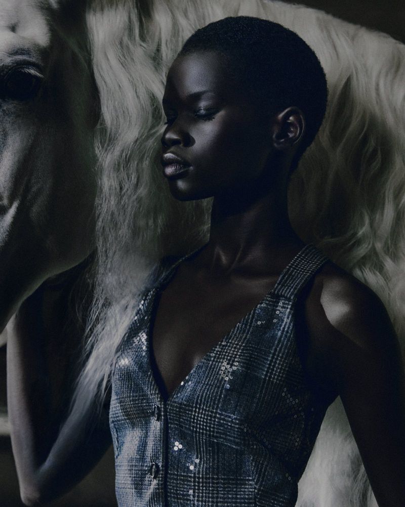 Portrait of a dark-skinned woman in a sleeveless vest with a white horse; the background is dark, and the lighting is soft.