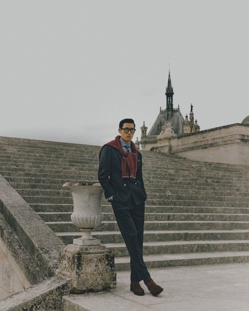 Adult man in a dark suit poses on stone steps in front of a historic building. The man is wearing glasses and brown boots.