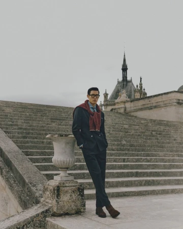 Adult man in a dark suit poses on stone steps in front of a historic building. The man is wearing glasses and brown boots.