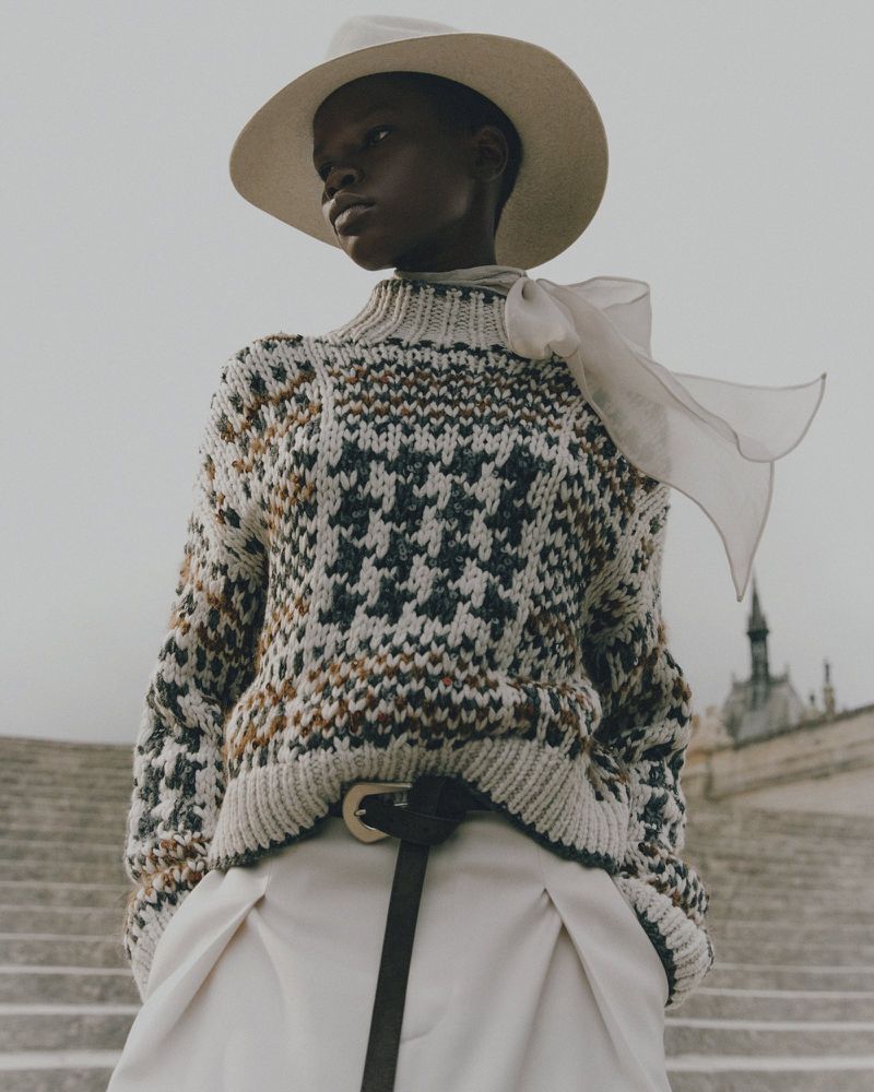 Woman wearing a houndstooth sweater, white pants, and a cream-colored hat poses on stone steps with a building in the background.