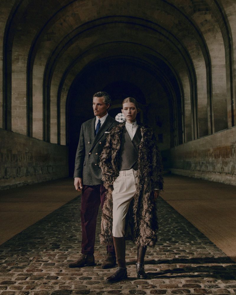 An adult man and woman stand side-by-side on a cobblestone path under an arched stone tunnel, both wearing tailored and layered clothing.