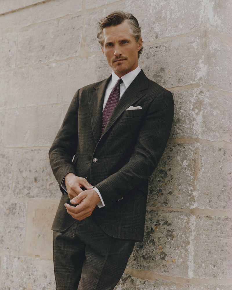 Adult male in a dark suit stands against a stone wall, hands clasped, looking at the camera in a medium, eye-level portrait.