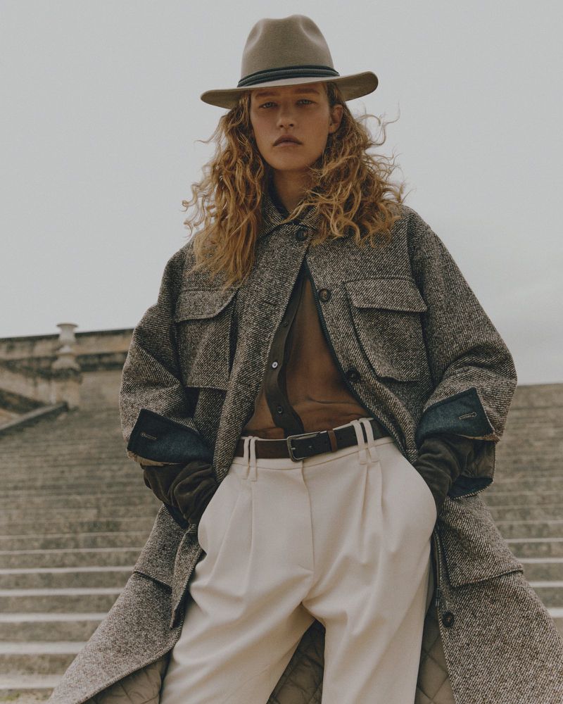 A woman with curly hair and a hat poses on stone steps wearing a jacket, blouse, belt, and pants. Cloudy skies in the background.