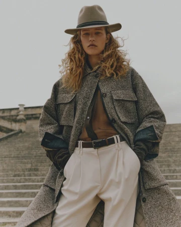 A woman with curly hair and a hat poses on stone steps wearing a jacket, blouse, belt, and pants. Cloudy skies in the background.