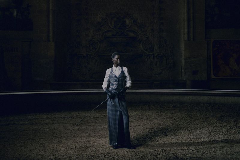 A solitary adult female stands centered in a dimly lit space wearing a vest, skirt, and riding gloves, holding a riding crop.