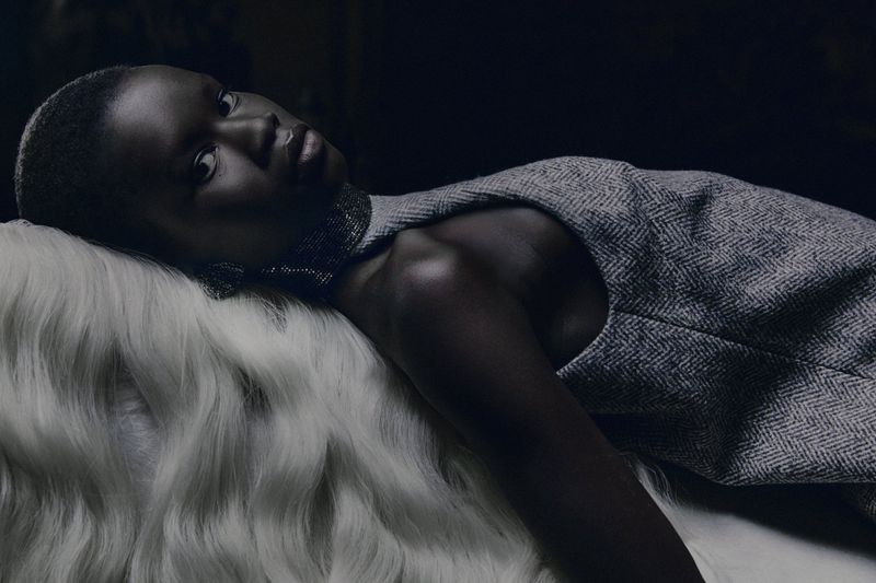 Dark-skinned adult female model in a gray dress, lying on white faux fur in a dimly lit environment, looking at the camera.