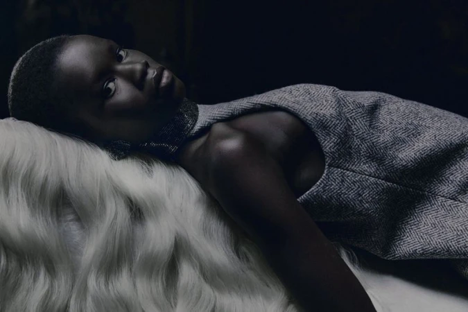 Dark-skinned adult female model in a gray dress, lying on white faux fur in a dimly lit environment, looking at the camera.