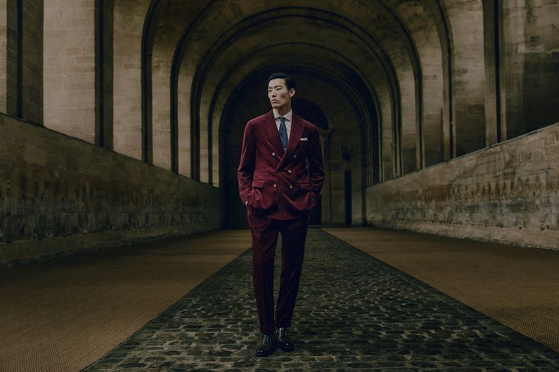 Asian man in a burgundy double-breasted suit stands in an arched stone hallway with cobblestone floors and diffused natural lighting.