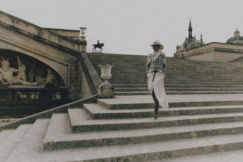 Woman in patterned sweater and skirt walks down grand stone steps of ornate building with equestrian statue in background.