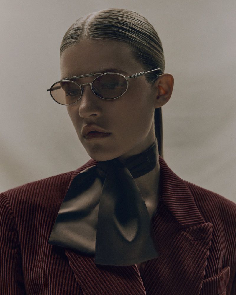 Fashionable portrait of a woman with hair slicked back, wearing a burgundy corduroy jacket, black necktie, and oval sunglasses.