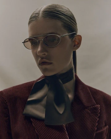Fashionable portrait of a woman with hair slicked back, wearing a burgundy corduroy jacket, black necktie, and oval sunglasses.
