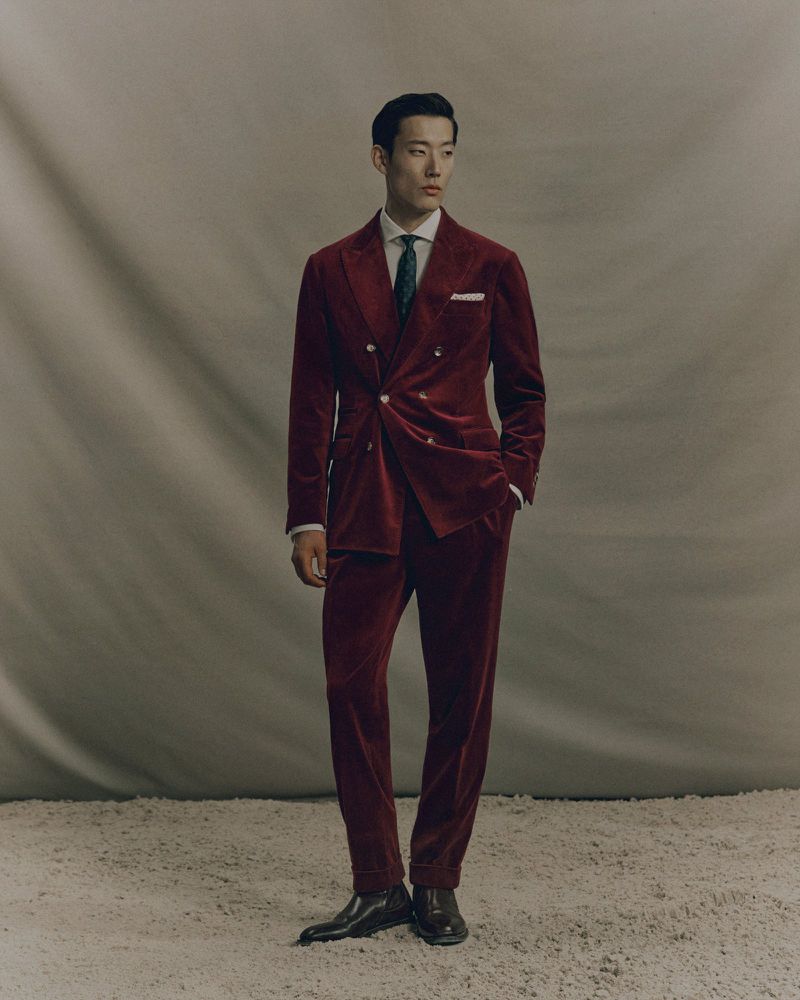 Asian man in a maroon velvet suit poses in a studio. The suit has a double-breasted jacket. He is wearing a white shirt and dark tie.